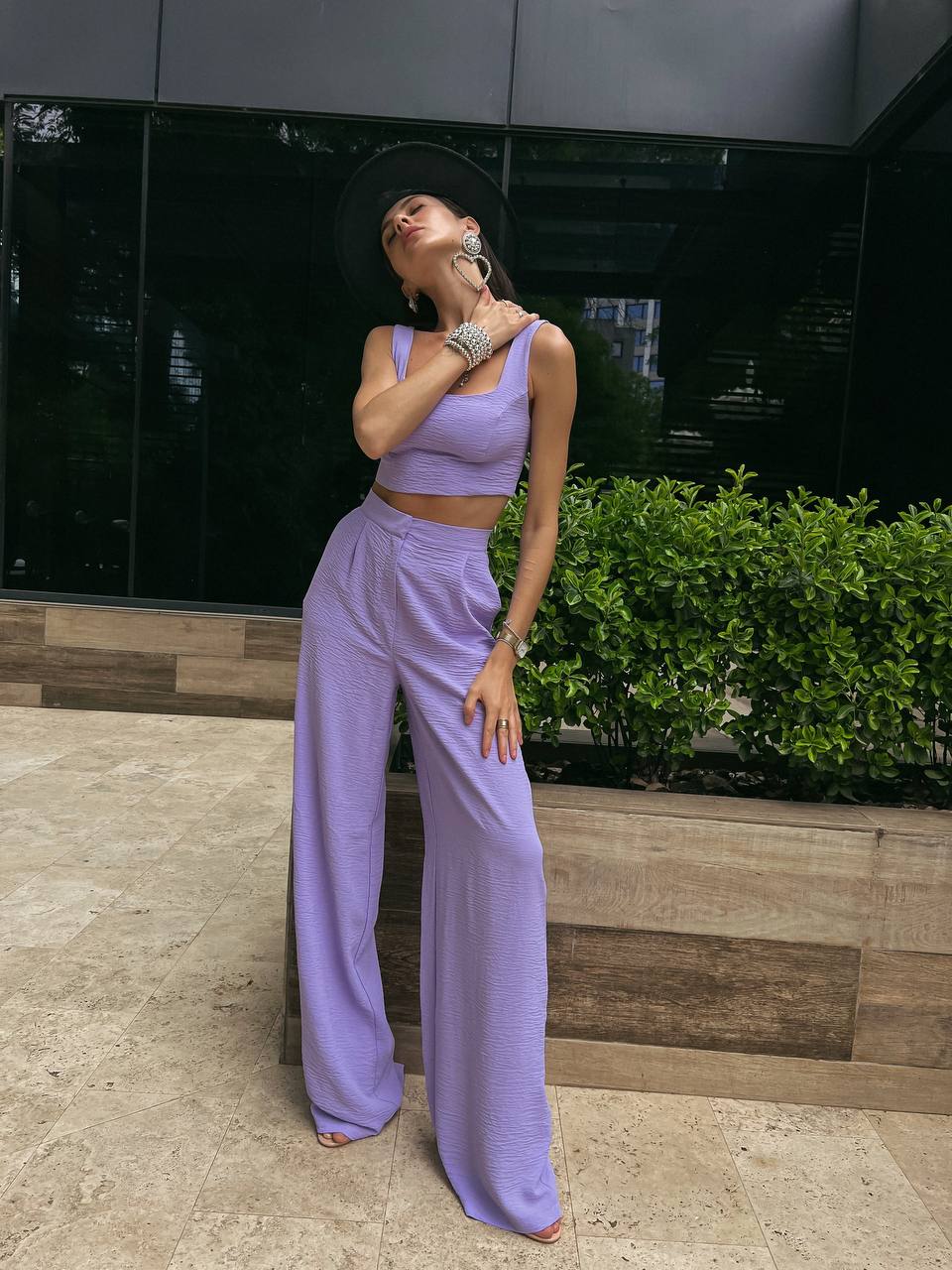Woman in a lavender outfit standing outdoors with a dark wall and greenery in the background