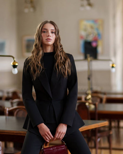 Woman in a black suit standing in an indoor setting with tables and chairs.