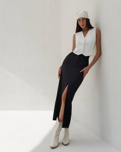 Woman wearing a white sleeveless top, black skirt with a slit, and white boots against a white background