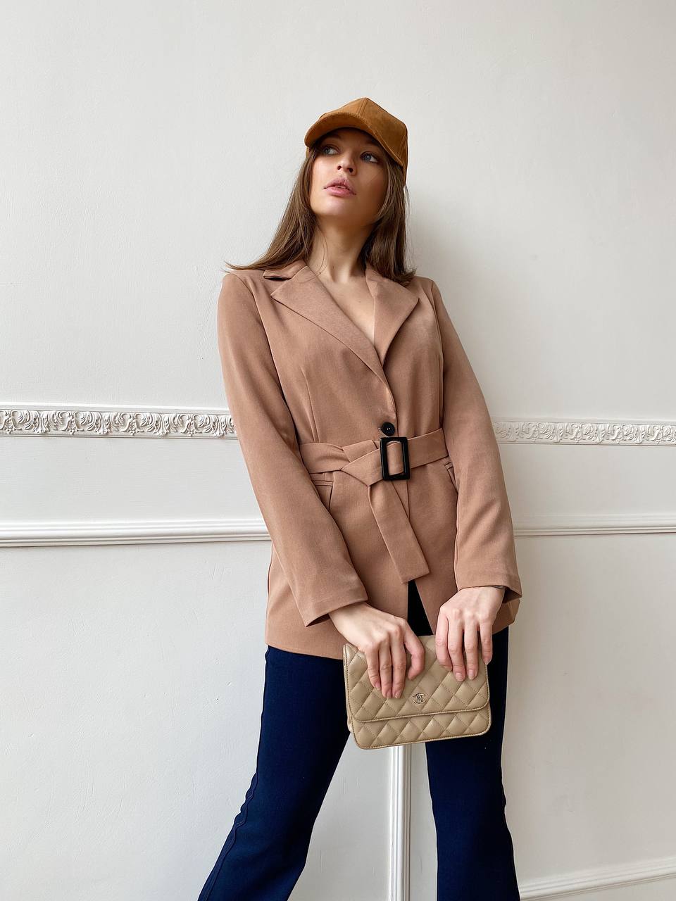 Woman wearing a beige blazer and cap, holding a beige clutch against a white wall.