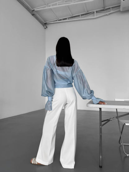 Person wearing a light blue sheer top and white pants in a minimalistic room.