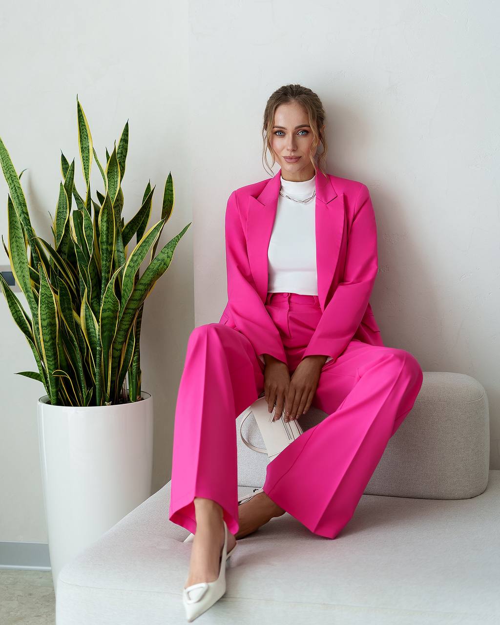 Woman in a bright pink suit sitting next to a potted plant indoors.