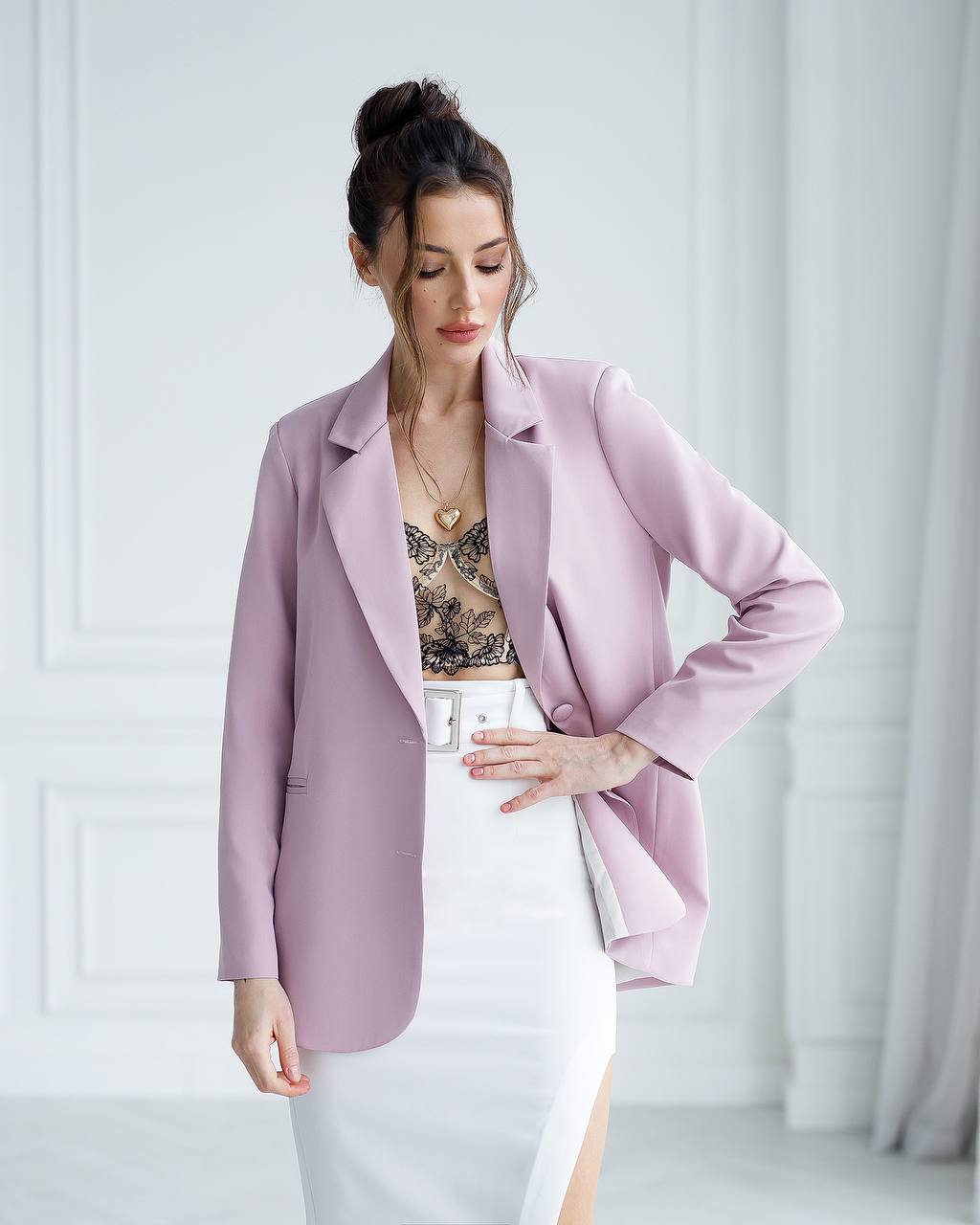 A woman wearing a lavender-purple blazer over a lace-trimmed top and a white skirt, standing in a bright, modern room with white paneled walls.