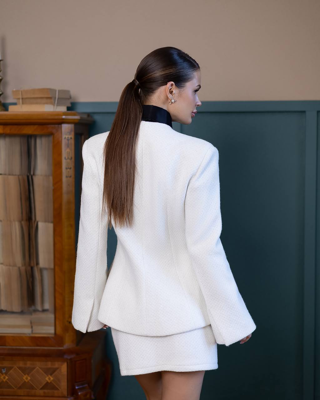 Woman in a white suit standing in a room with a wooden cabinet and green wall.