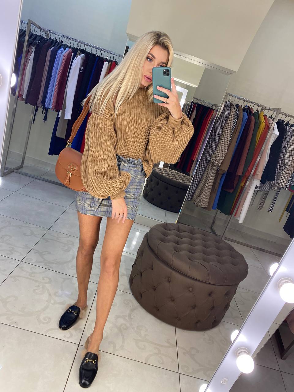 Woman taking a mirror selfie in a clothing store, wearing a brown ribbed sweater and plaid skirt.