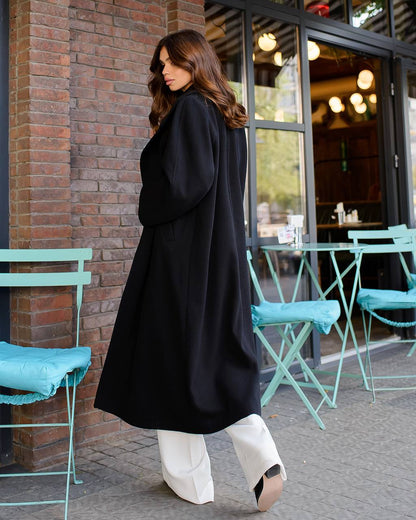 Woman in a black coat walking past a cafe with teal chairs.