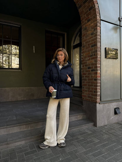 Woman in a blue puffer jacket and beige pants standing outside a building.