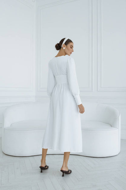 Woman wearing a white dress standing in a minimalistic room with a white sofa.