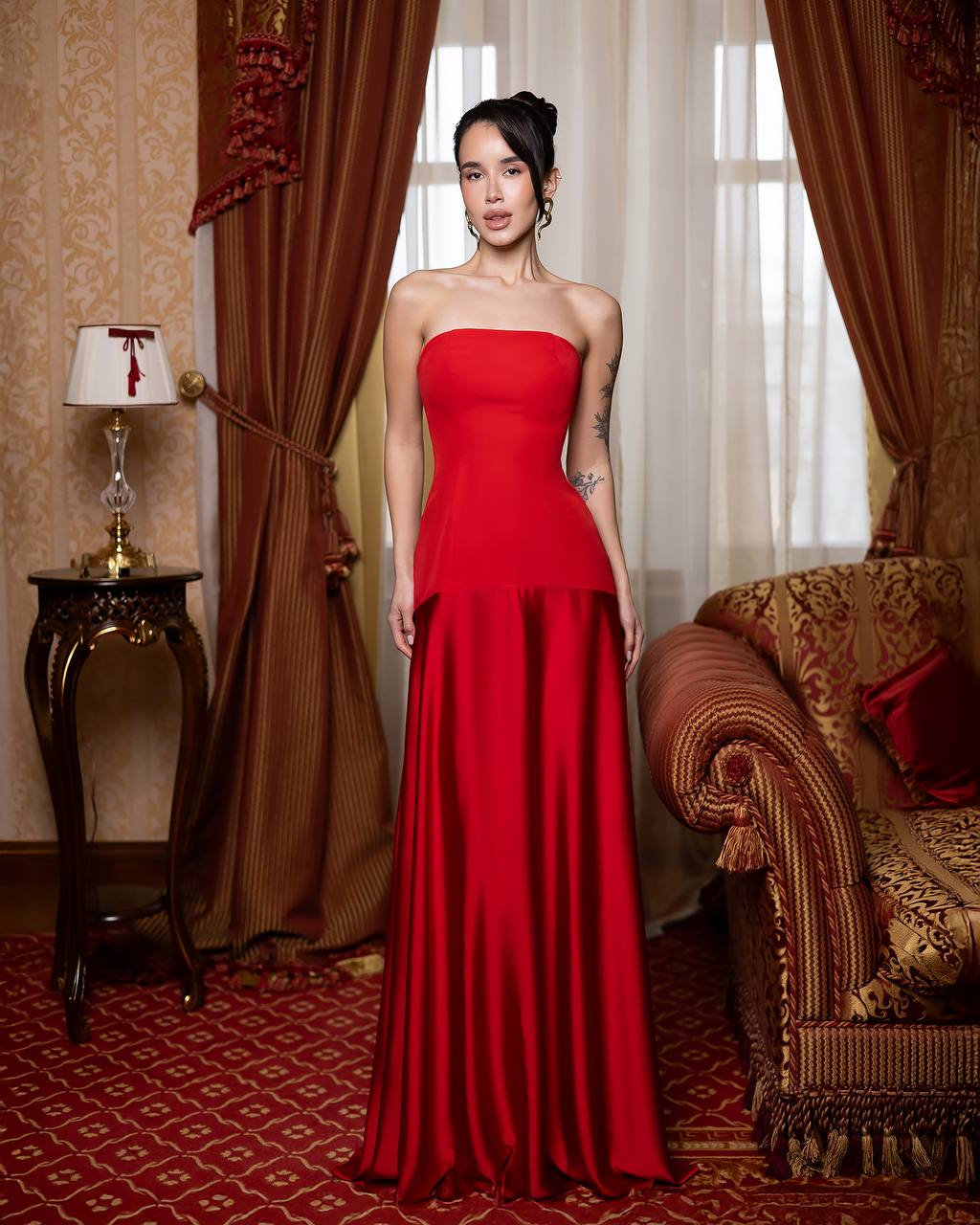 Woman in a red strapless gown standing in an ornate room with curtains and a sofa.