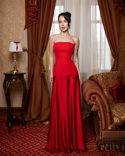 Woman in a red strapless gown standing in an ornate room with curtains and a sofa.