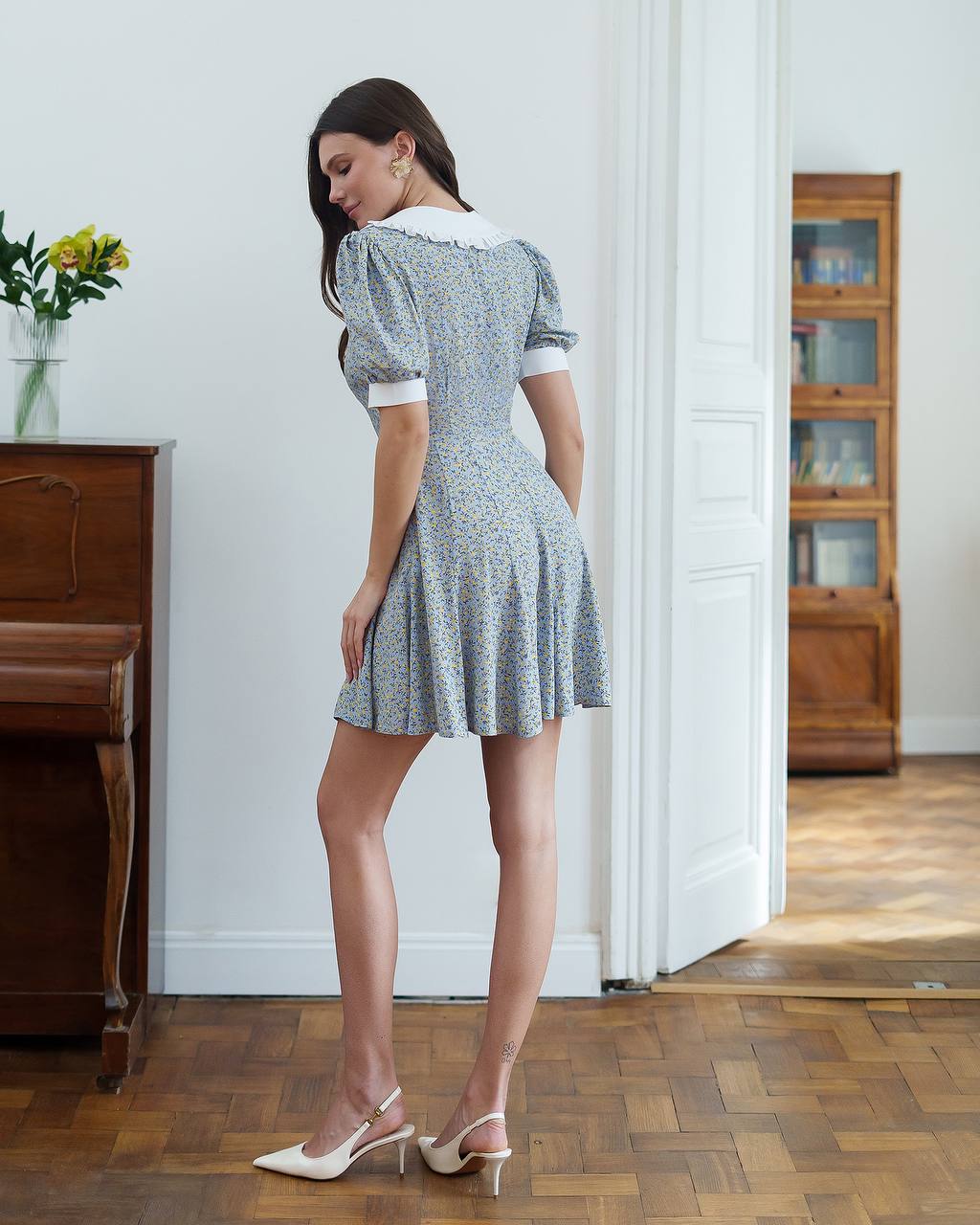 Woman in a patterned dress standing in a room with wooden floors and furniture.