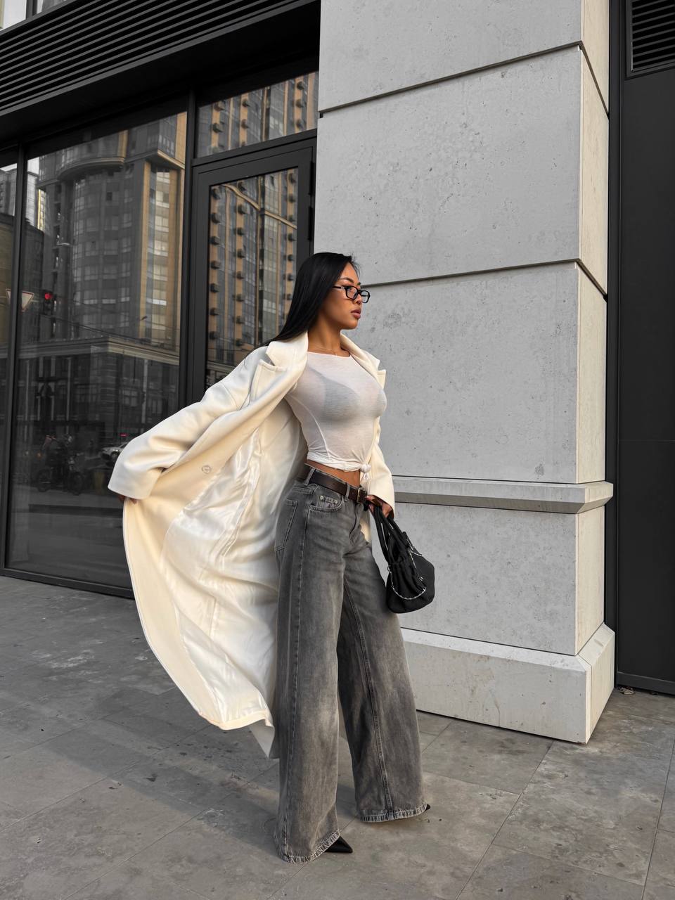 Woman in a white coat and gray pants standing against a building.