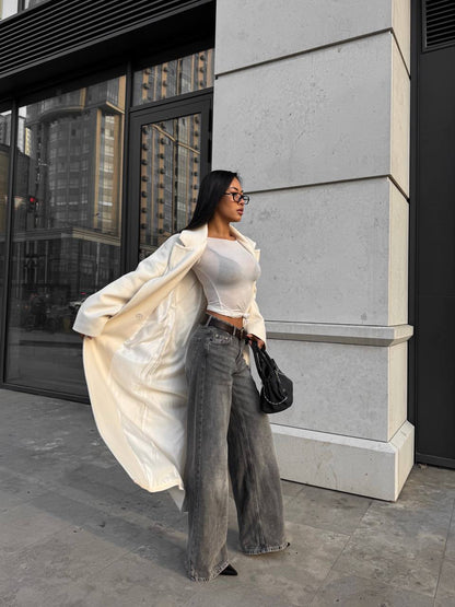 Woman in a white coat and gray pants standing against a building.