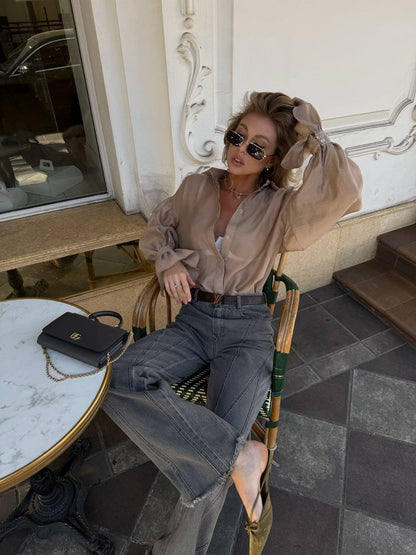 Woman sitting outdoors wearing a beige blouse and blue jeans, with sunglasses on.