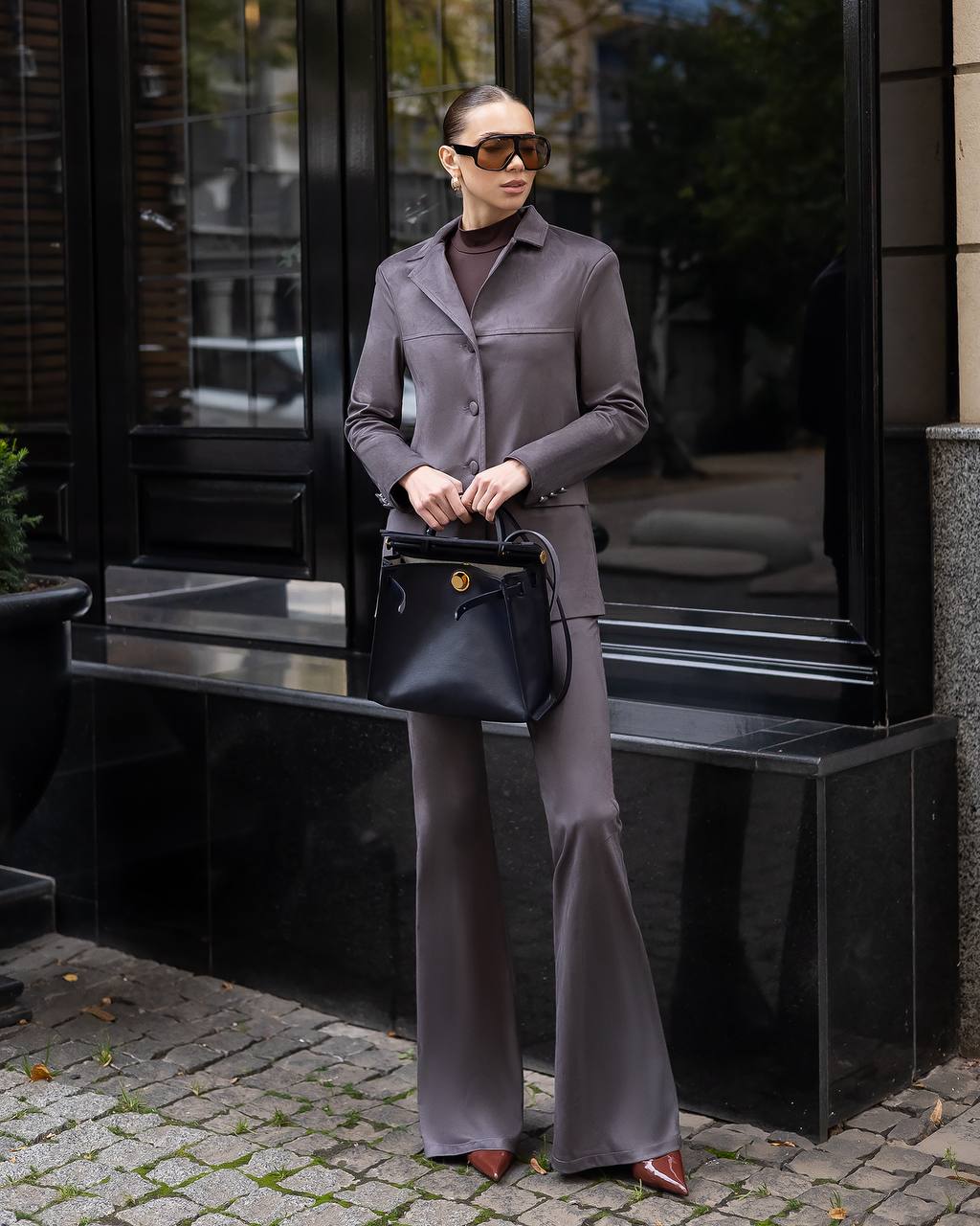 Woman in a gray outfit holding a black handbag on a city street.