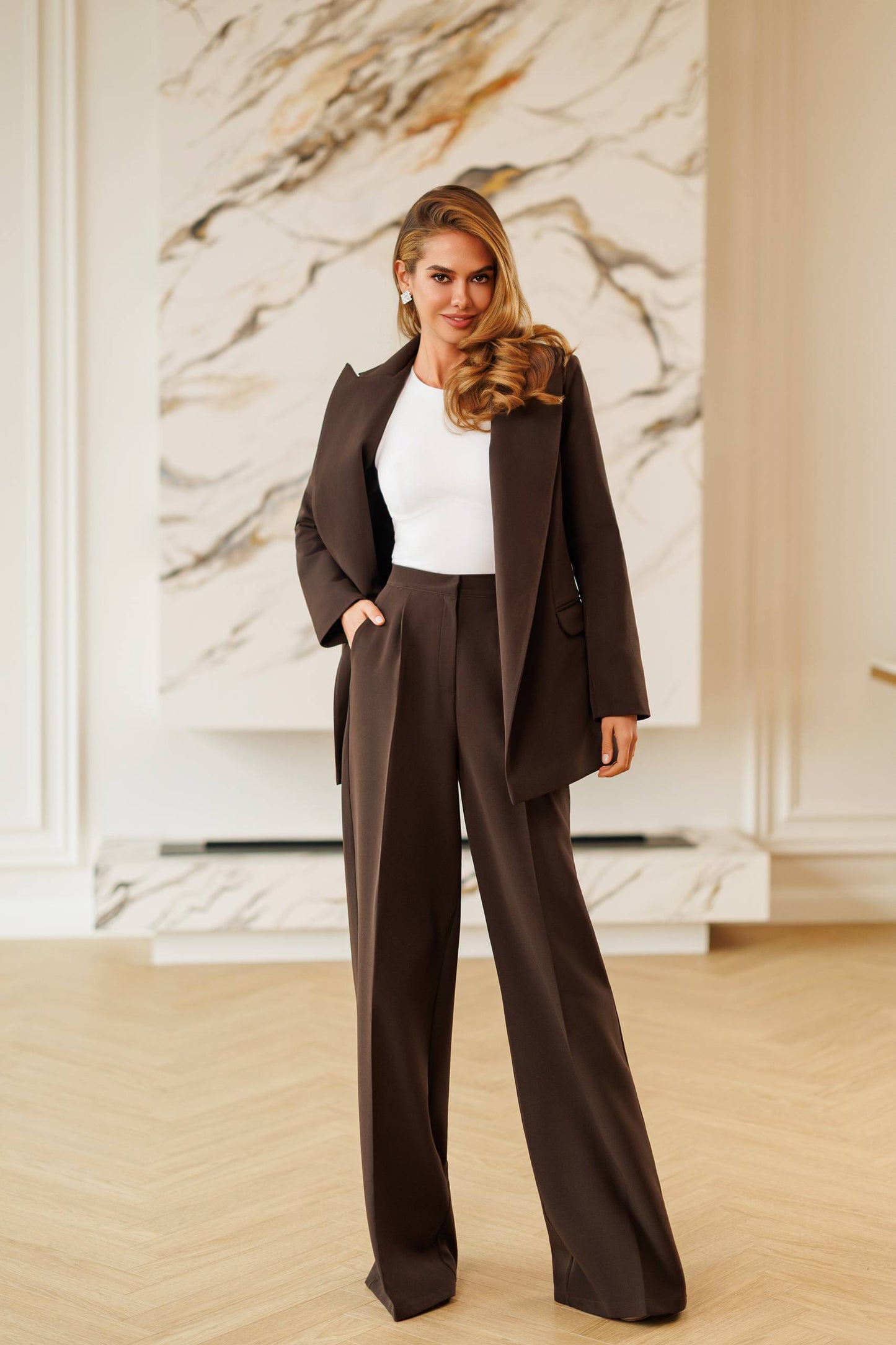 A woman modeling a dark brown tailored suit with a blazer and wide-leg trousers, styled with a white top, standing in a bright room with light wood floors and a marble wall art piece.