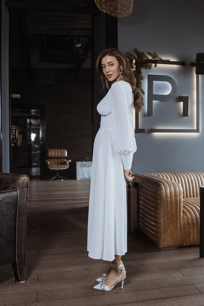 Woman in a white dress standing in a stylish interior setting with a 'P' logo on the wall.