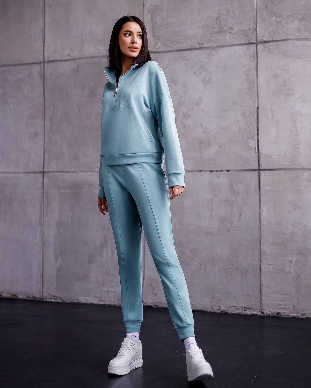 Woman wearing a light blue tracksuit against a gray concrete wall.
