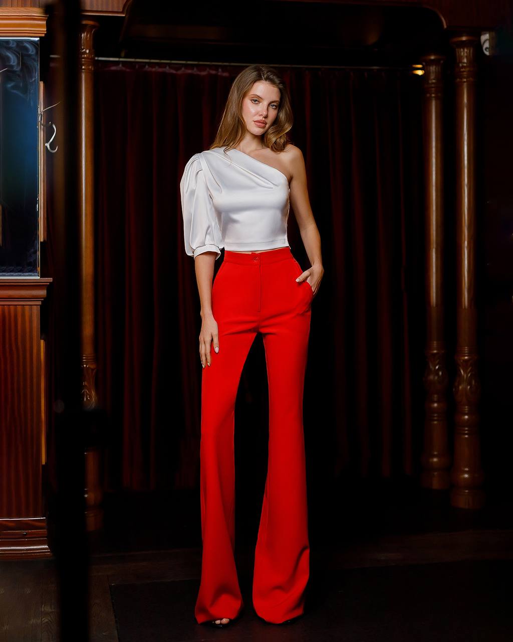 Woman wearing a white top and red pants standing in a room with wooden paneling.