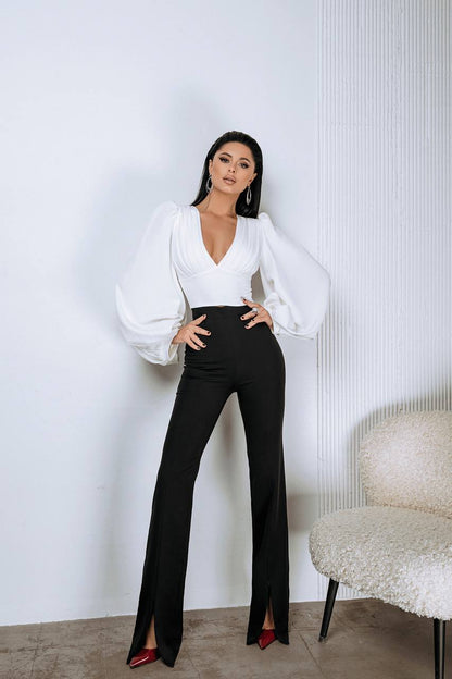 Woman in a white blouse and black pants posing against a white wall.