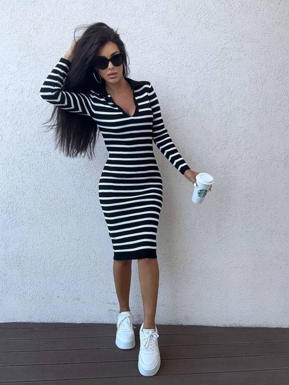 Woman wearing a black and white striped dress with white sneakers against a plain wall.