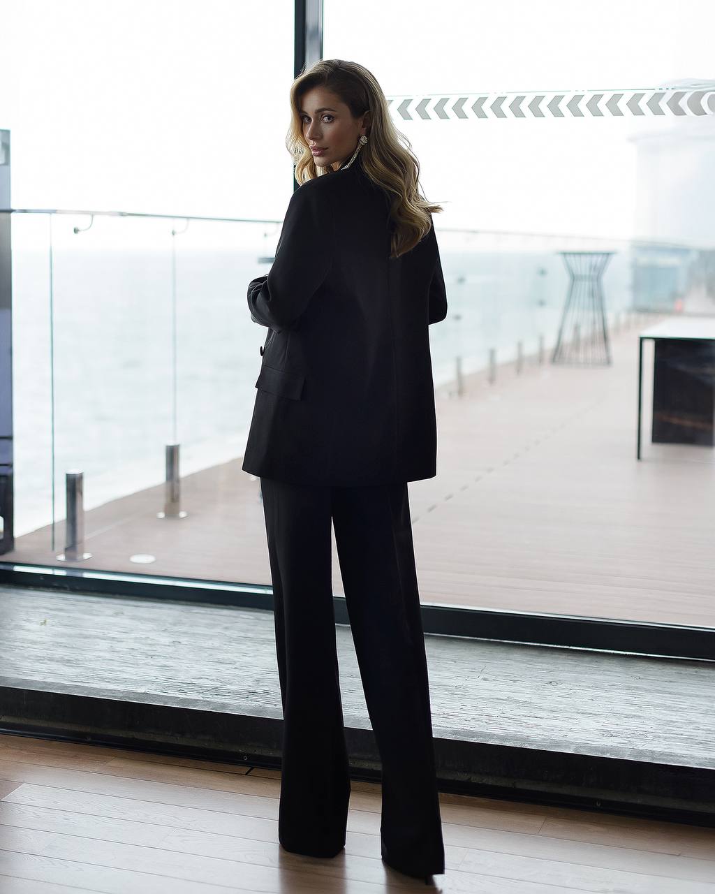Woman in a black suit standing in a modern indoor setting with large windows.