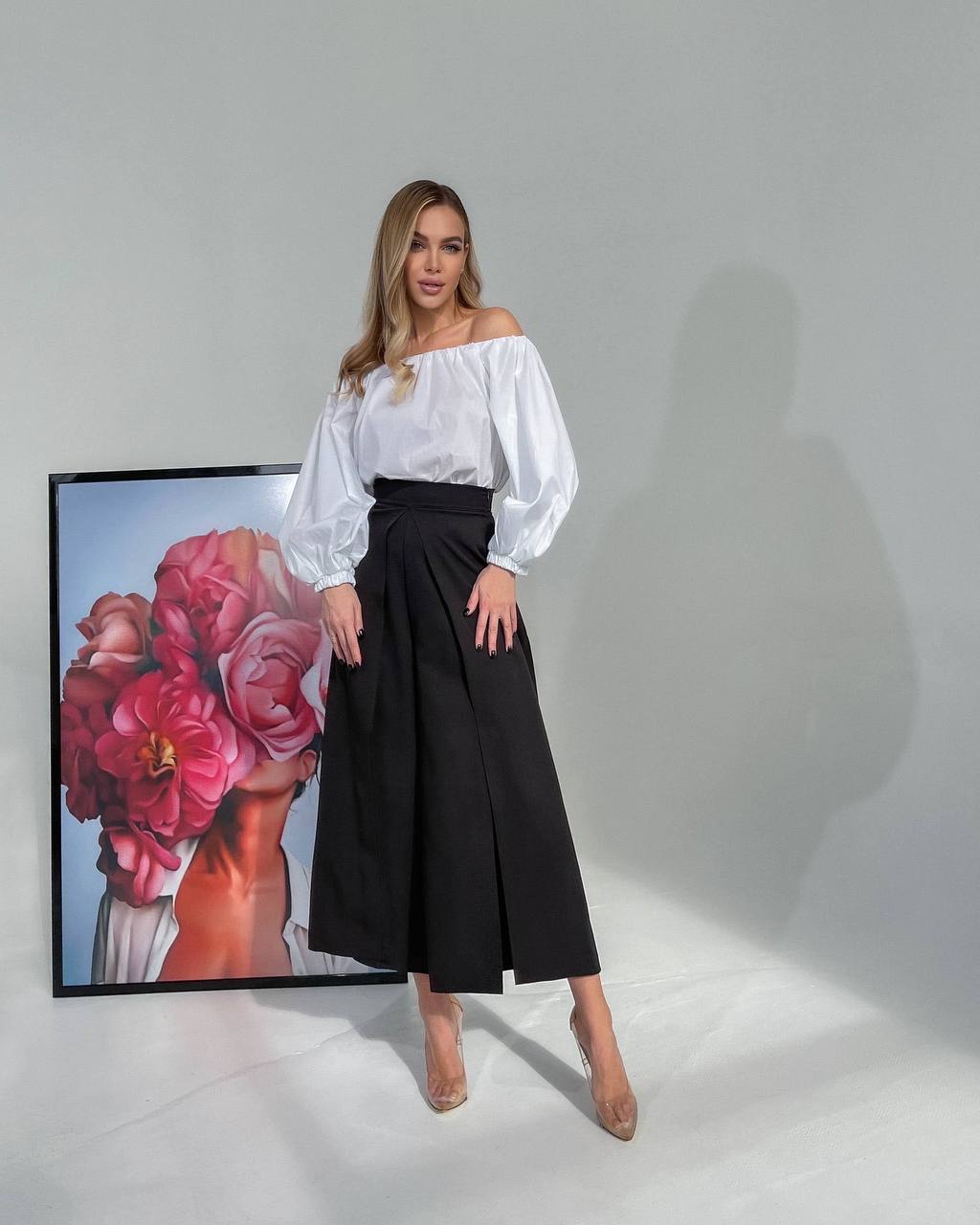 Woman in a white blouse and black skirt standing next to a framed artwork of flowers.
