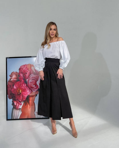Woman in a white blouse and black skirt standing next to a framed artwork of flowers.