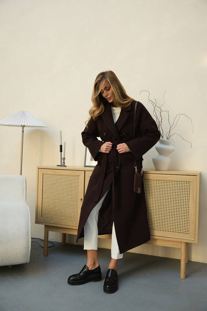 Woman in a brown coat standing in a room with light-colored walls and furniture.