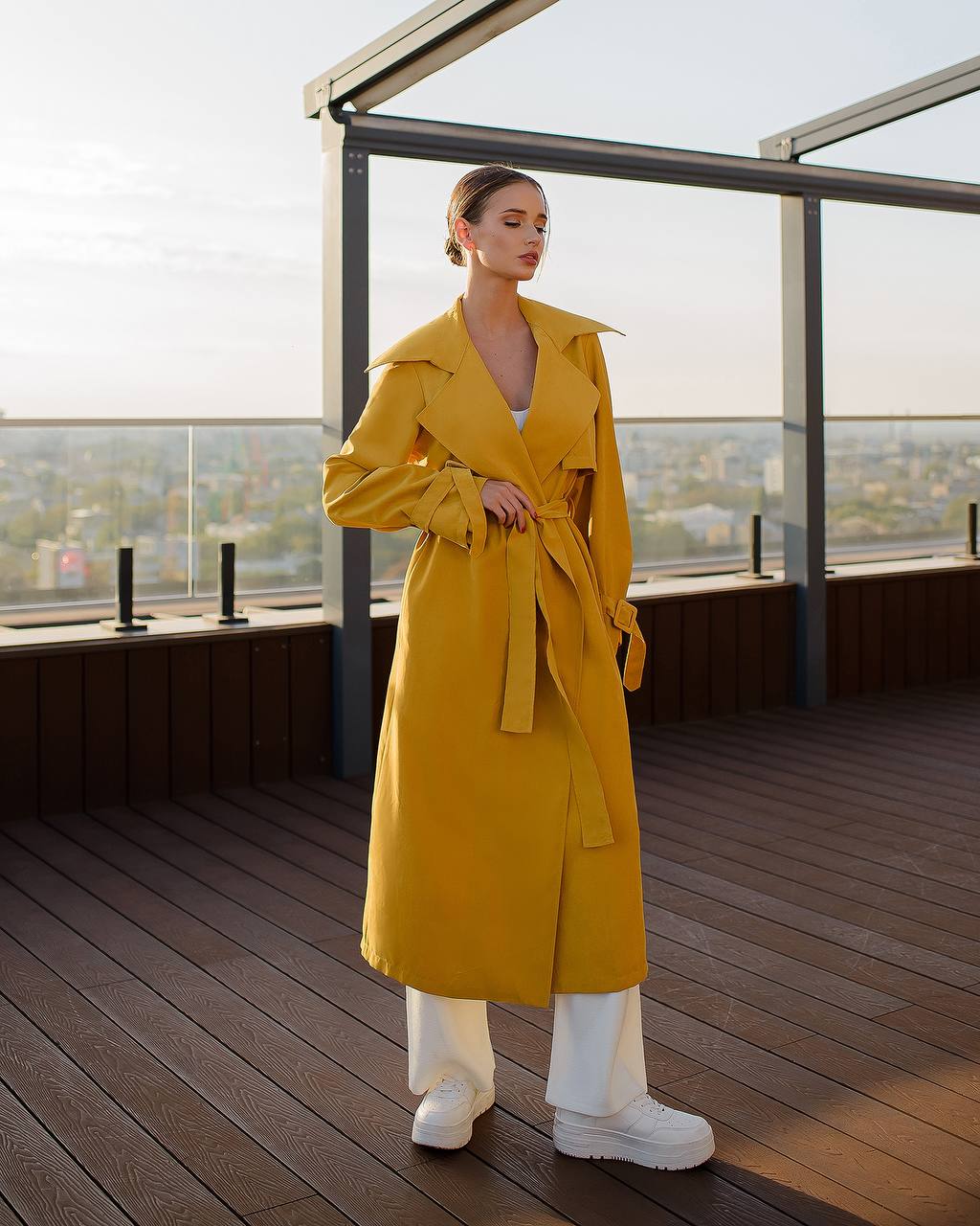 Woman in a yellow trench coat standing on a rooftop with a cityscape view.
