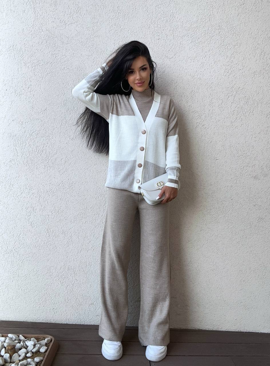 Woman wearing a two-tone cardigan and beige pants against a plain wall.