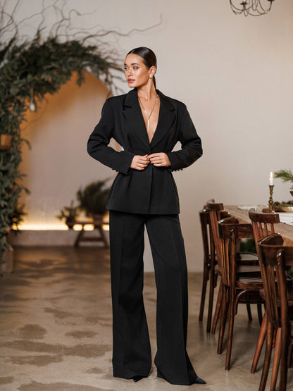 Woman in a black suit standing in a room with wooden furniture and decorative plants.