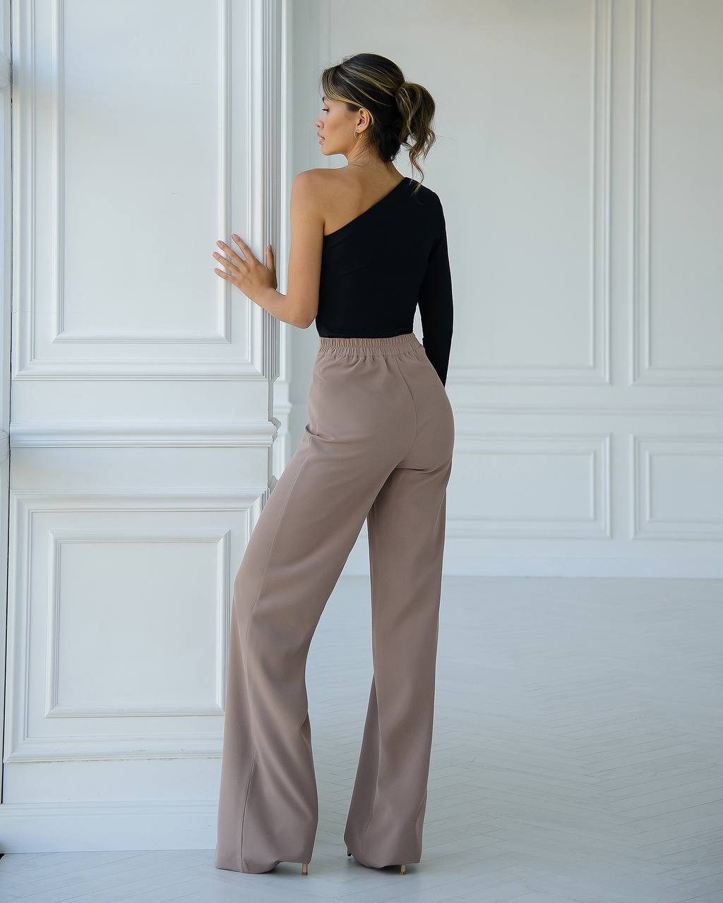 Woman wearing a black top and beige pants against a white paneled wall.