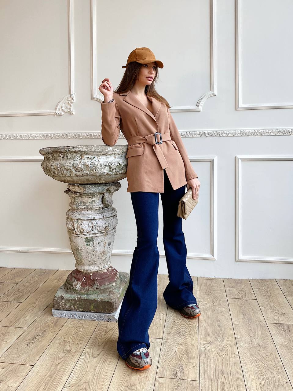 Woman in a beige coat and blue pants standing in an elegant interior setting.