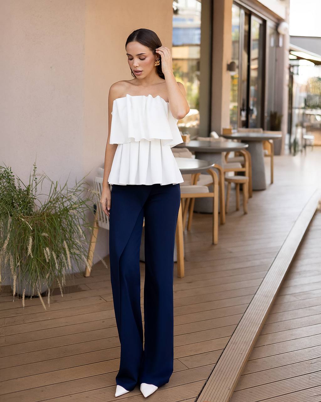 A woman stands on a wooden outdoor patio wearing a white off-the-shoulder ruffled top and navy wide-leg trousers, with white pointed shoes.