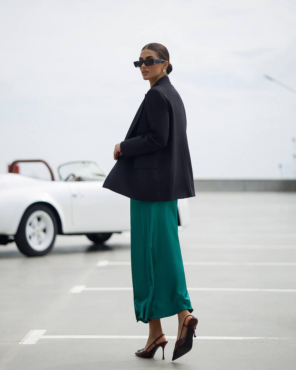 Woman in a black blazer and green skirt standing in a parking lot with a white car in the background.