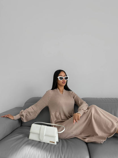 Woman in a beige dress sitting on a gray couch with a white handbag, wearing sunglasses.