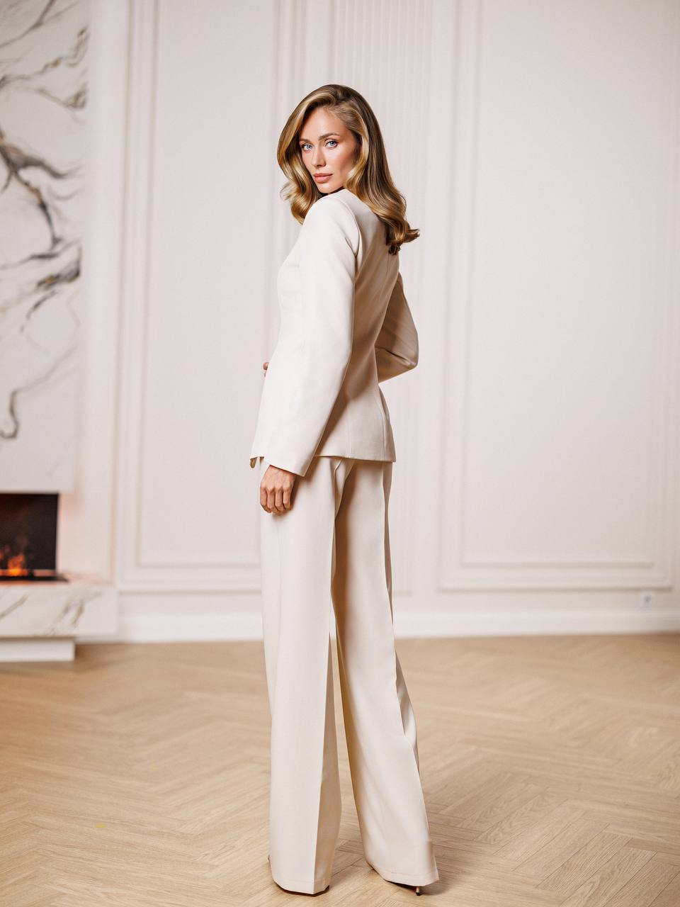 Woman in a beige suit standing in a room with a fireplace and marble wall.