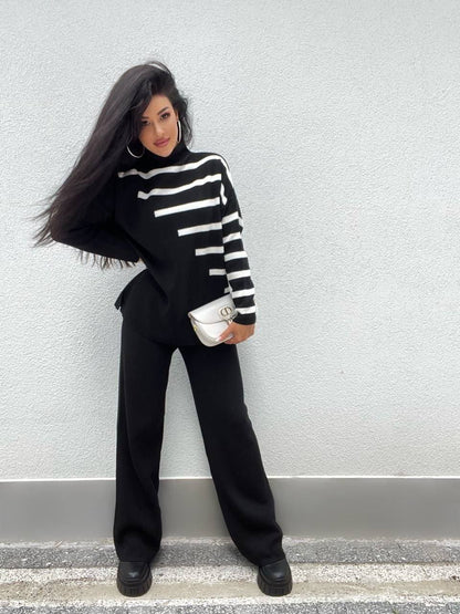 Woman wearing a black and white striped sweater and black pants against a light gray wall.