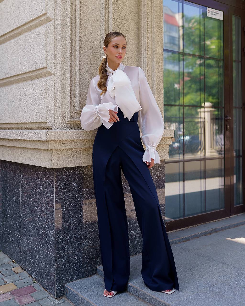 Woman in a white blouse and navy pants standing against a building.