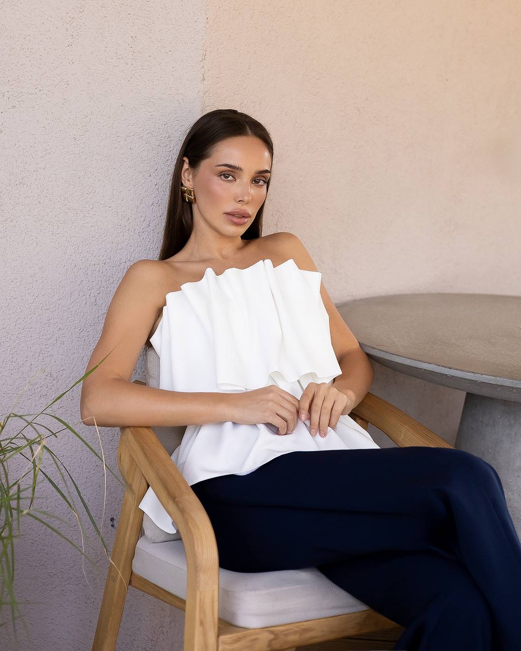 A woman sits in a wooden chair wearing a white strapless top with dramatic ruffle detailing and dark navy pants, posed against a textured beige wall.