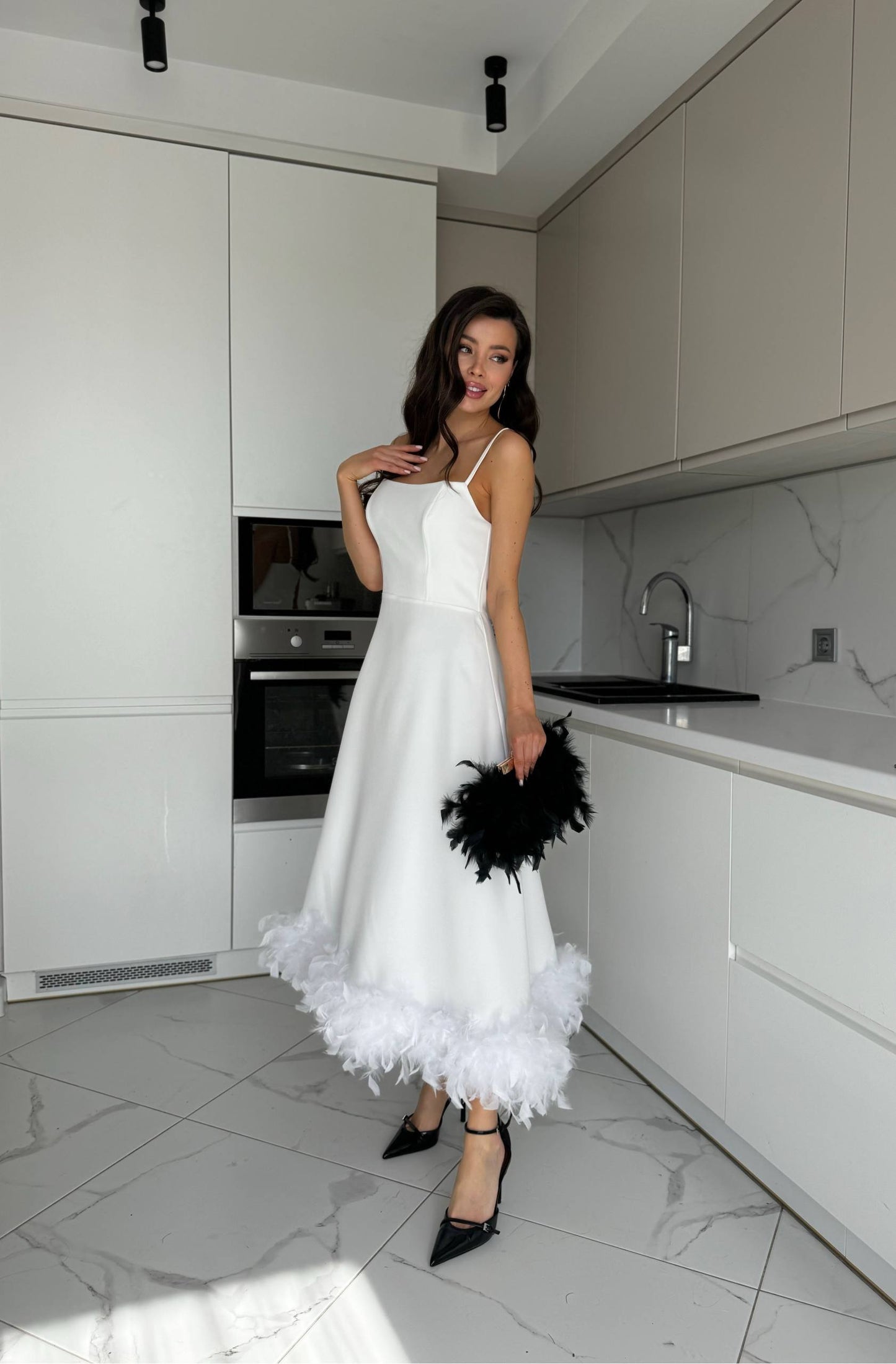 Woman in a white dress with black feathers standing in a modern kitchen.