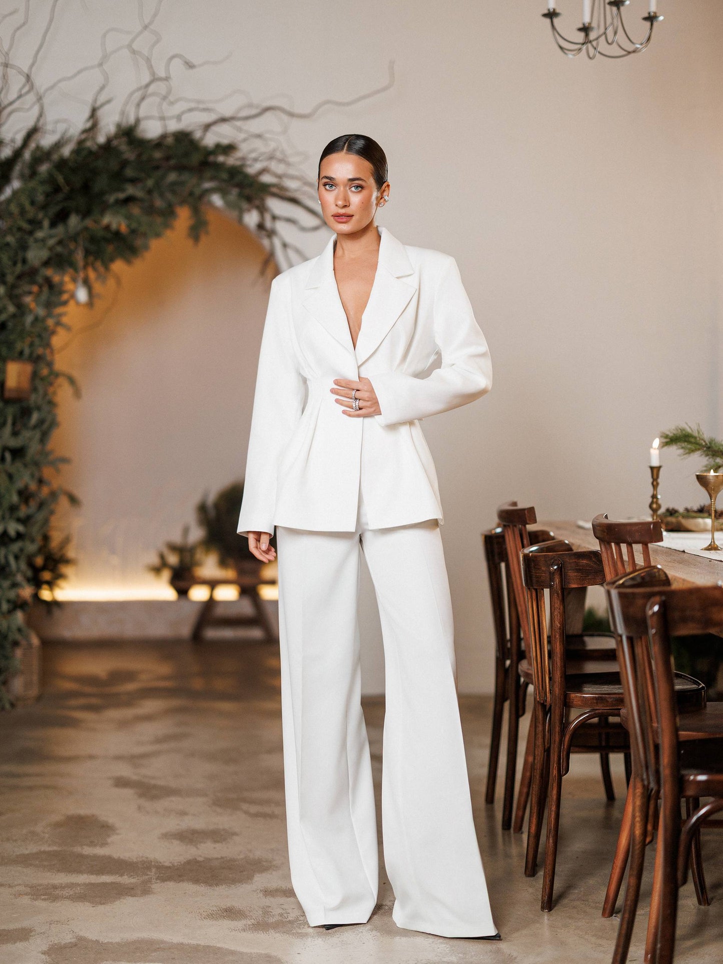 Woman in a white suit standing in a room with wooden furniture and decorative plants.