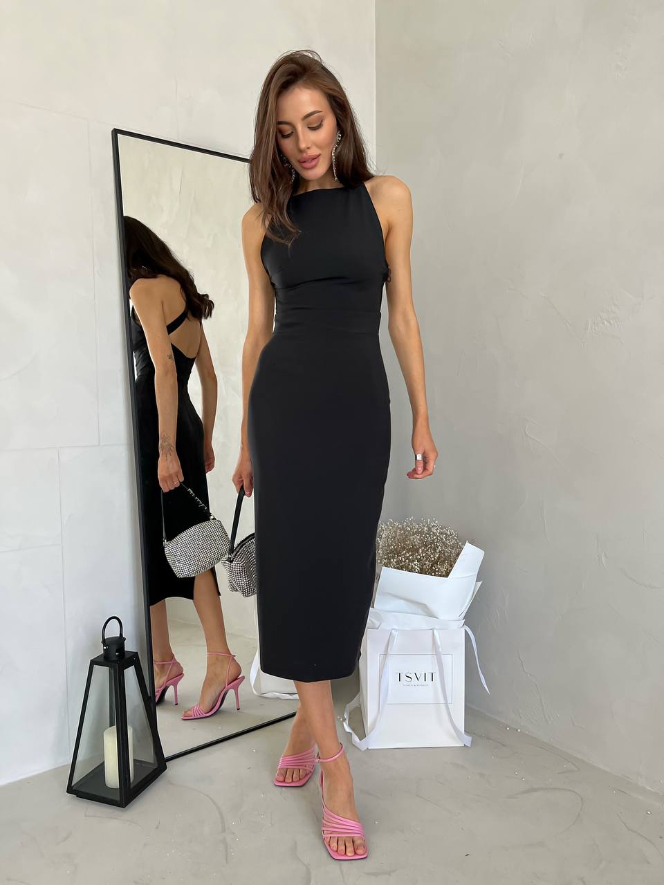 Woman in a black sleeveless dress standing in front of a mirror, with a white bag and lantern on the floor.