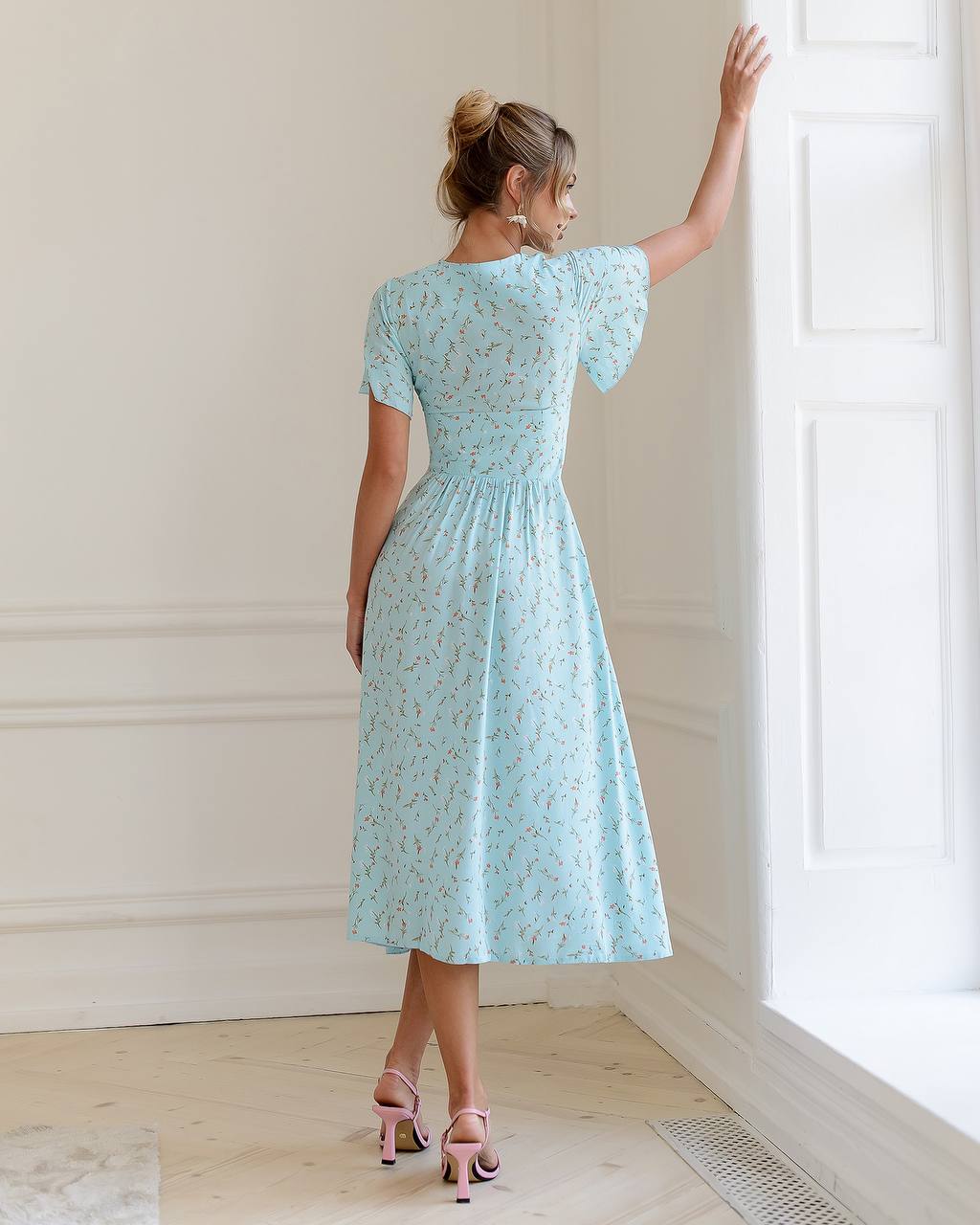 Woman in a light blue floral dress standing in a bright room with white walls.