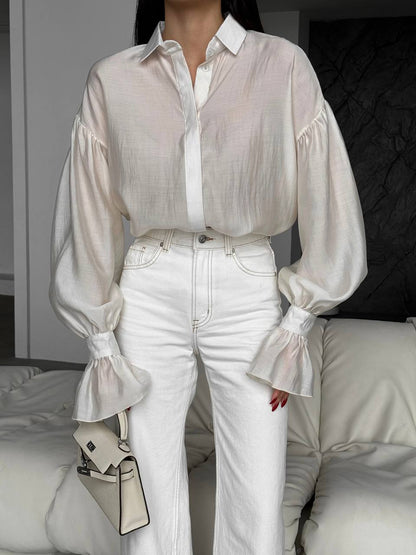 Person wearing a sheer white blouse with long sleeves and white pants, holding a beige handbag.