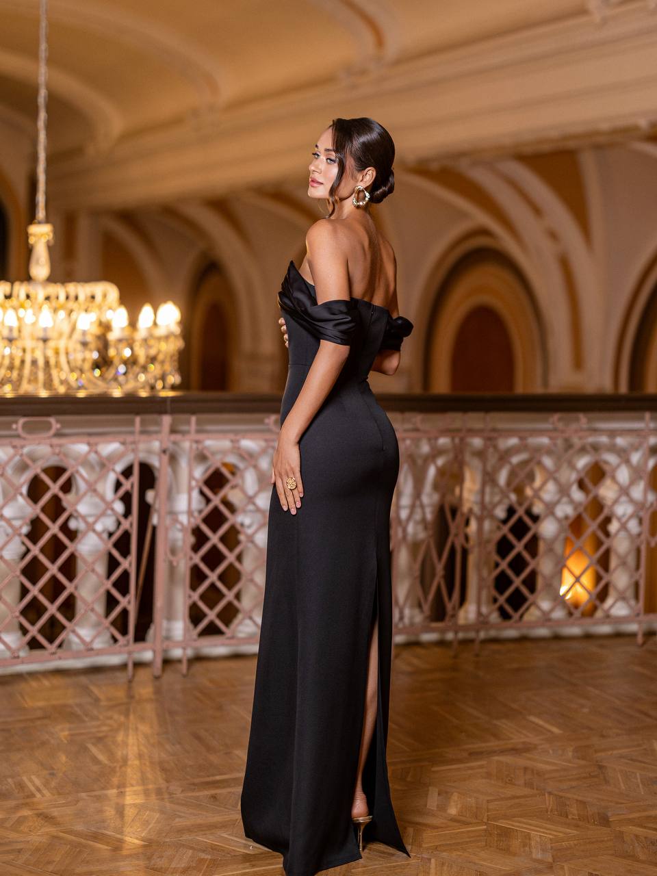 Woman in a black evening gown standing in an elegant indoor setting with chandeliers.