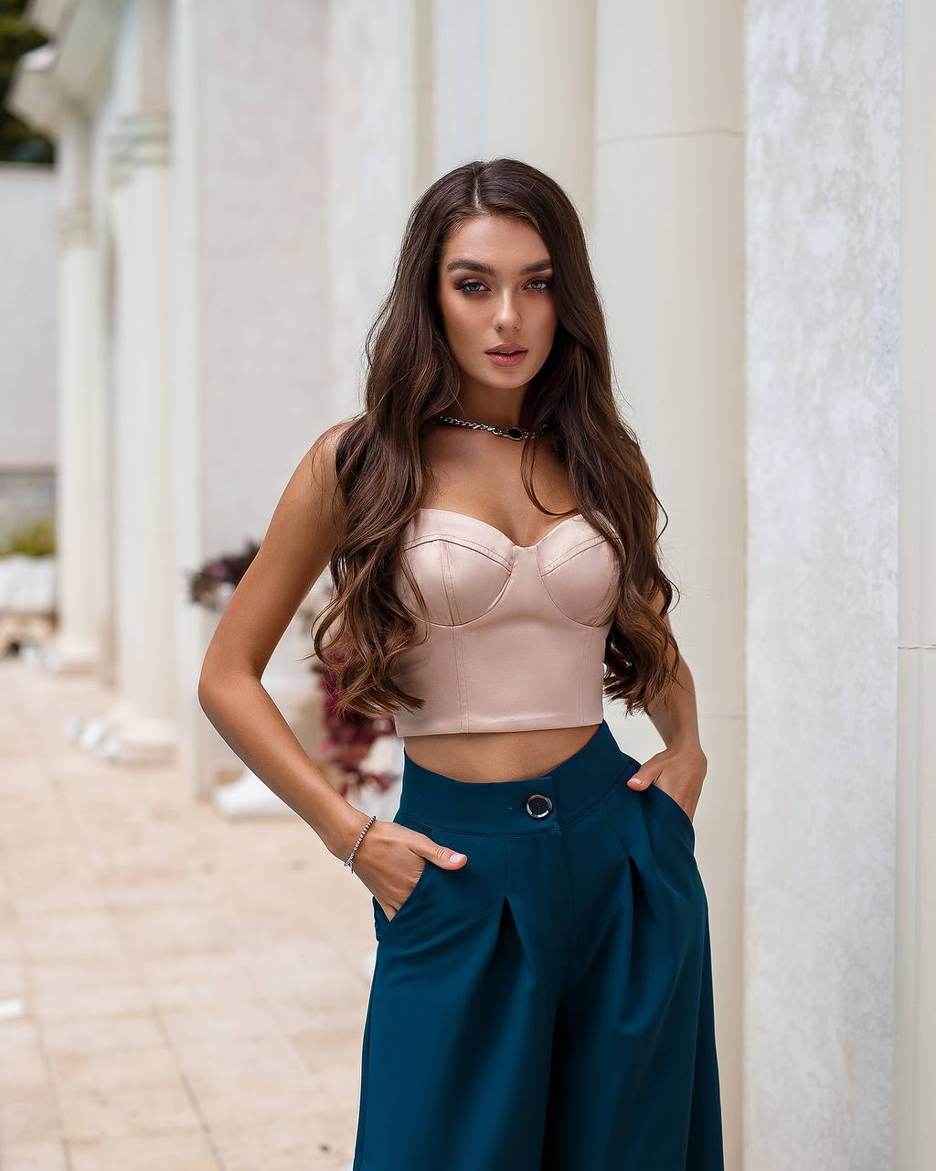 Woman wearing a pink top and teal pants standing against a white architectural background