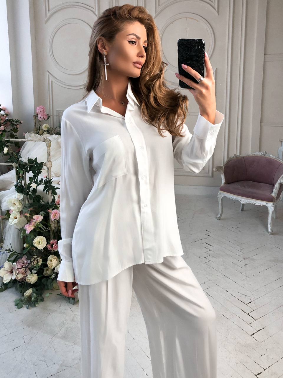 Woman in a white outfit taking a selfie in a stylish room with flowers and furniture.
