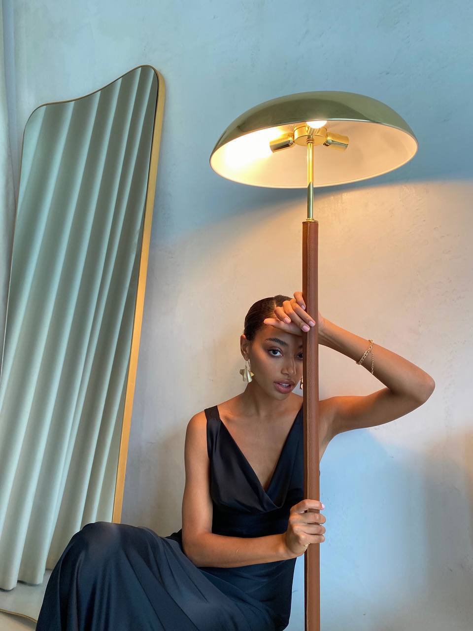 Woman in a dark blue dress holding a tall floor lamp against a light blue wall.